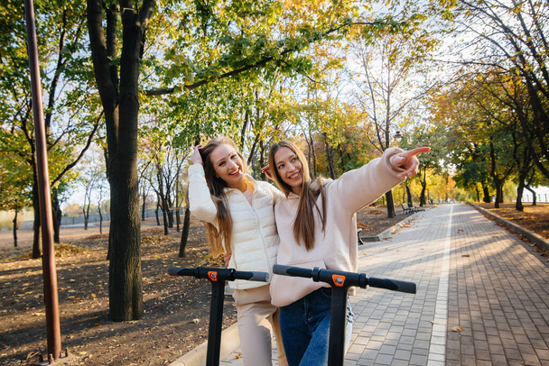 Two young beautiful girls ride electric scooters in the Park on a warm autumn day - Zdjęcie, obraz