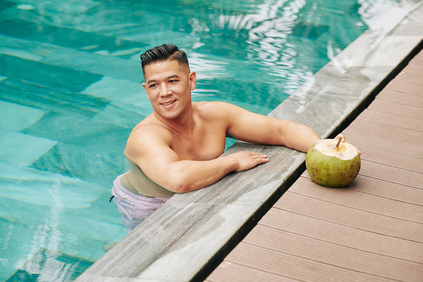 Alegre hombre de mediana edad de pie en la piscina del hotel spa y beber cóctel de coco delicioso dulce - Foto, Imagen