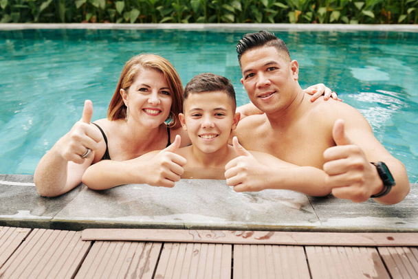 Neşeli baba, anne ve oğul yüzme havuzunda baş parmağını kaldırıyor. - Fotoğraf, Görsel
