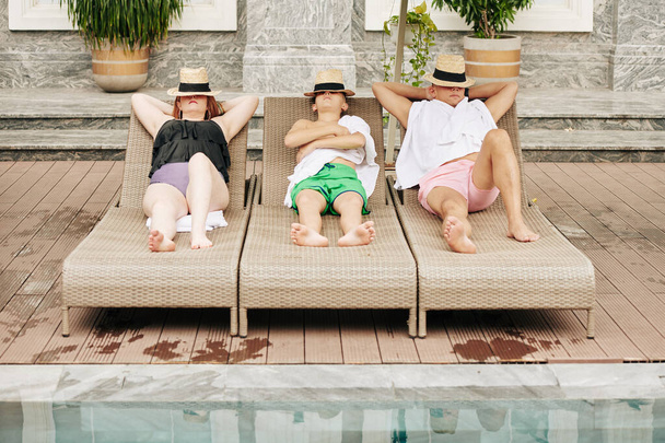 Vater, Mutter und Sohn bedecken Gesichter mit Hut beim Sonnenbaden auf Liegestühlen am Pool - Foto, Bild