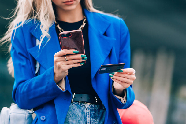 Happy fashion blonde shopper wearing a blue jacket paying on line with a credit card and a smart phone on the street - 写真・画像