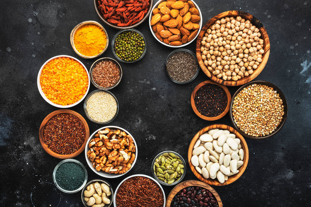 Selection of superfoods, legumes, cereals, nuts, seeds in bowls on black background. Superfood as chia, spirulina, beans, goji berries, quinoa, turmeric and other. Copy space, top view - Фото, изображение