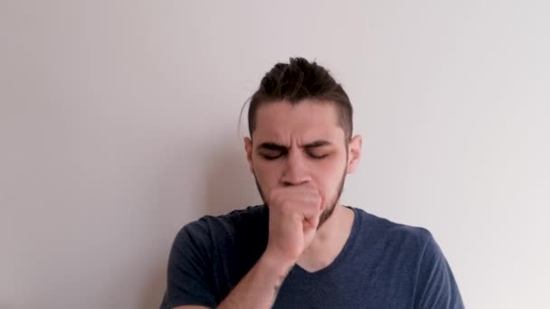 Young handsome Caucasian man coughs in palm of his hand, blows his nose in paper napkin, and feels very ill. Man is ill and coughs violently snivels against background of white wall. - Footage, Video