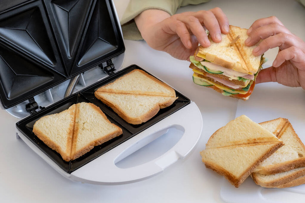 Ręce trzymać gotowane świeże kanapki z serem, bekonem i warzywami w pobliżu toster na białym stole. - Zdjęcie, obraz