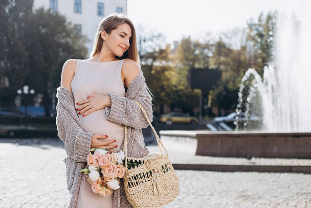 Pretty pregnant woman with flowers walking on the street. Beautiful blonde girl with brown eyes. Professional make-up. Happy and carefree pregnancy. Autumn street. - Foto, imagen