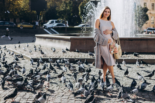 Beautiful pregnant mother stands near the pigeons in a sunny summer park. Walking on the street. Professional make-up. Happy and carefree pregnancy. Summer street. - Fotoğraf, Görsel