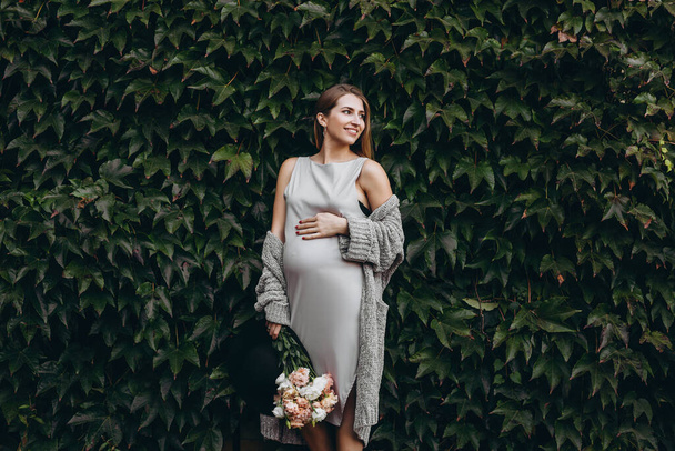 A young pregnant girl holds a bouquet of flowers and touches her baby. Happiness. Smile. Nature. Wall of green leaves. - Фото, изображение