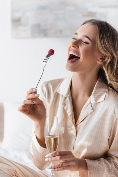 Positive woman in pajama holding champagne and raspberry on fork in bedroom  - Photo, Image