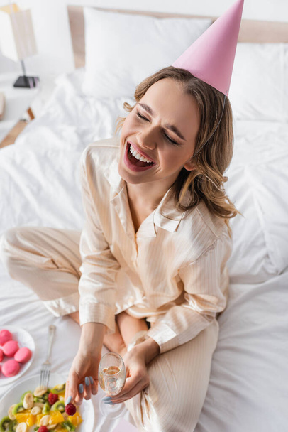 Cheerful woman in party cap holding champagne and raspberry on bed  - Foto, Imagem