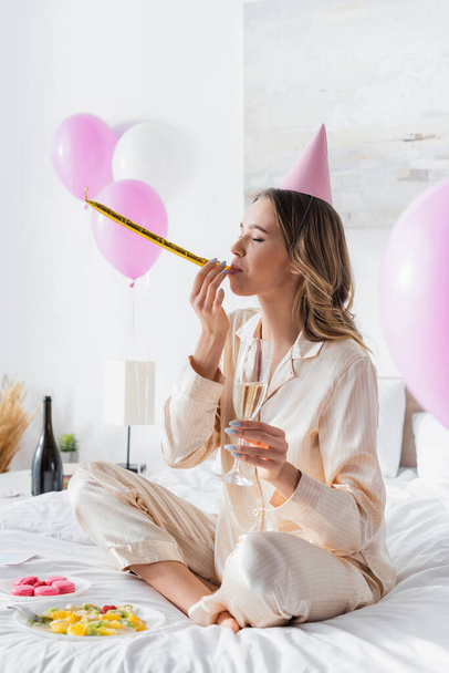 Woman blowing party horn and holding champagne near macaroons and fruit salad on bed  - Foto, Imagem