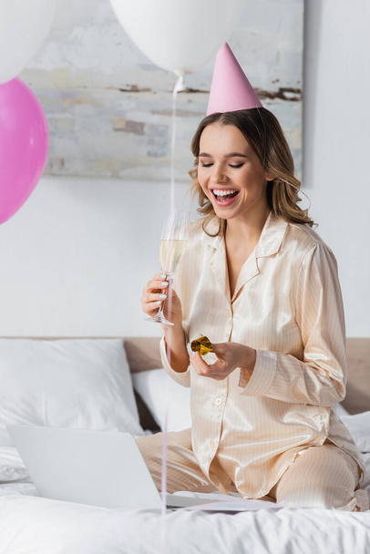 Smiling woman in pajama holding champagne and party horn near laptop on bed  - Foto, imagen