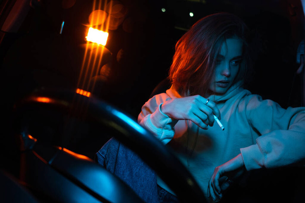 Sad girl in neon light in a car with a cigarette. - Photo, image