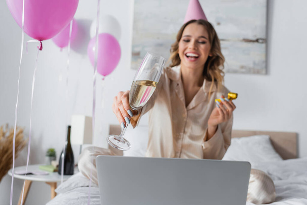 Champagne in hand of blurred woman celebrating birthday near laptop on bed  - Valokuva, kuva