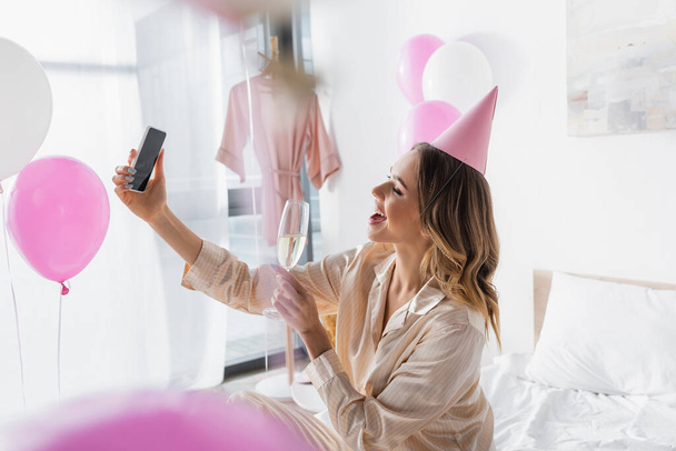 Side view of cheerful woman with champagne taking selfie on smartphone in bedroom  - Fotó, kép