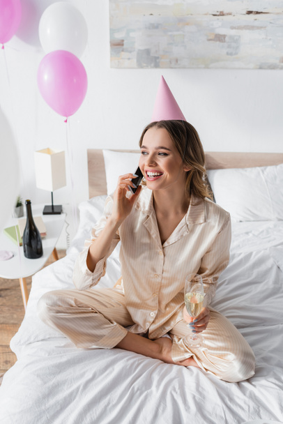 Smiling woman with champagne and party cap talking on smartphone on bed  - 写真・画像