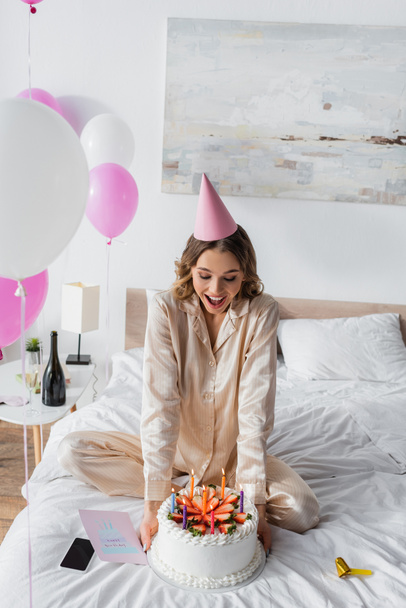 Возбужденная женщина в роге для вечеринок, держит торт на день рождения рядом с открыткой и смартфоном  - Фото, изображение