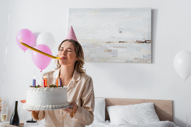 Mujer soplando cuerno de fiesta y celebración de pastel de cumpleaños en la cama  - Foto, Imagen