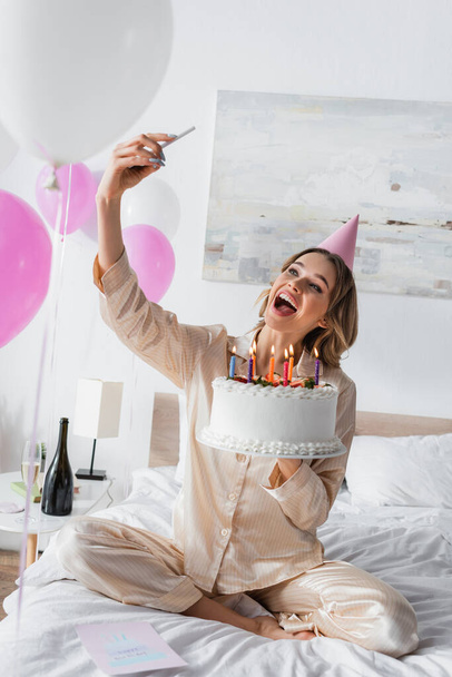 Excited woman in party cap taking selfie with smartphone and holding birthday cake  - Foto, imagen