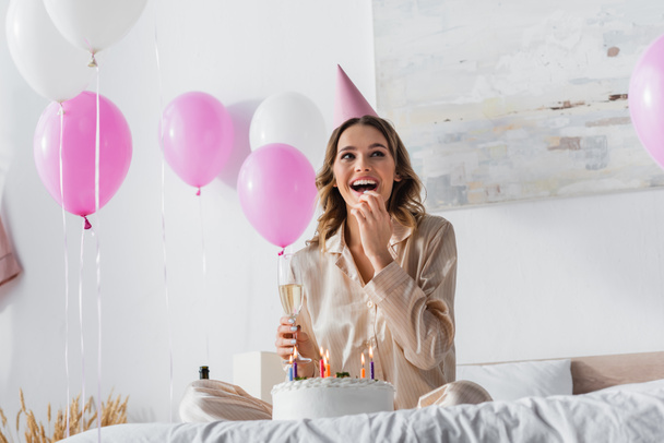Cheerful woman holding glass of champagne near birthday cake with candles on bed  - 写真・画像