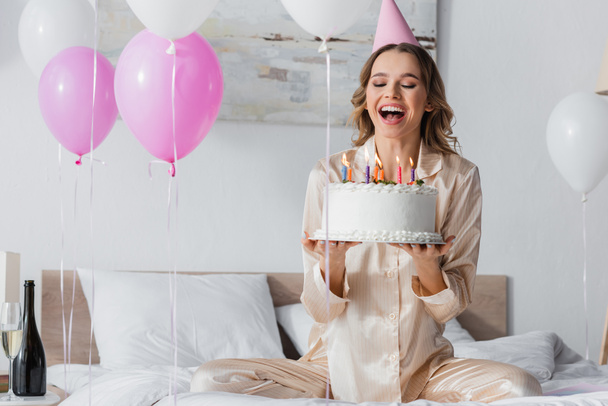 Mujer feliz con gorra de fiesta sosteniendo pastel de cumpleaños cerca de globos y champán en el dormitorio  - Foto, Imagen
