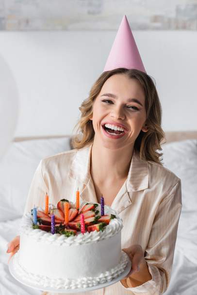 Smiling woman in party cap looking at camera while holding birthday cake at home  - Fotografie, Obrázek