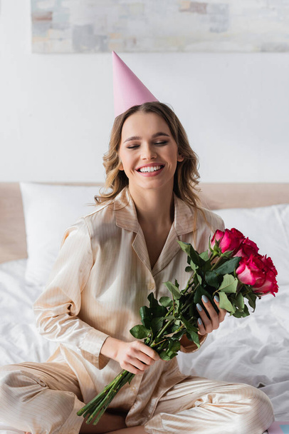 Femme souriante en pyjama et bonnet de fête tenant des roses sur le lit  - Photo, image
