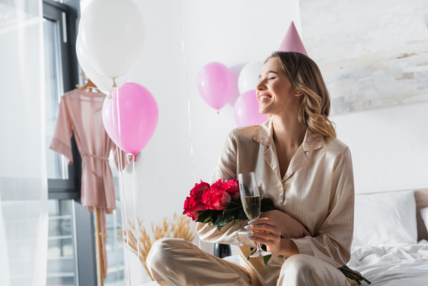 Uśmiechnięta kobieta trzyma bukiet i szampana podczas urodzin w domu  - Zdjęcie, obraz