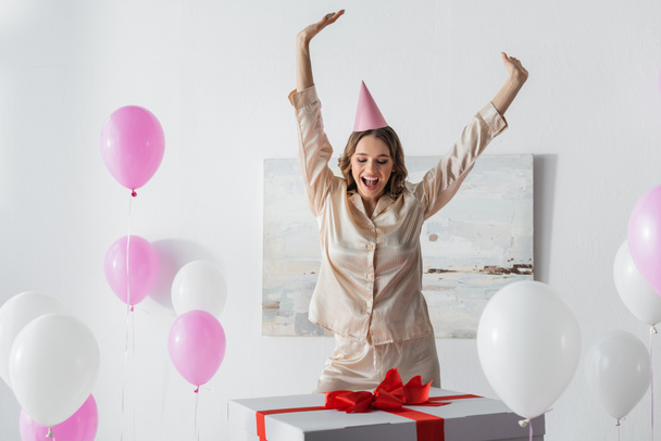 Positive Frau im Pyjama neben riesigem Geschenk und Luftballons zu Hause  - Foto, Bild