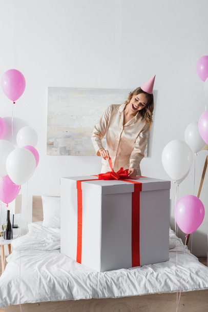 Mujer alegre en gorra de fiesta sosteniendo arco de regalo enorme cerca de globos en el dormitorio  - Foto, imagen