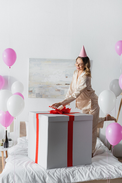 Young woman in pajama and party cap standing near big present on bed and balloons  - Photo, Image