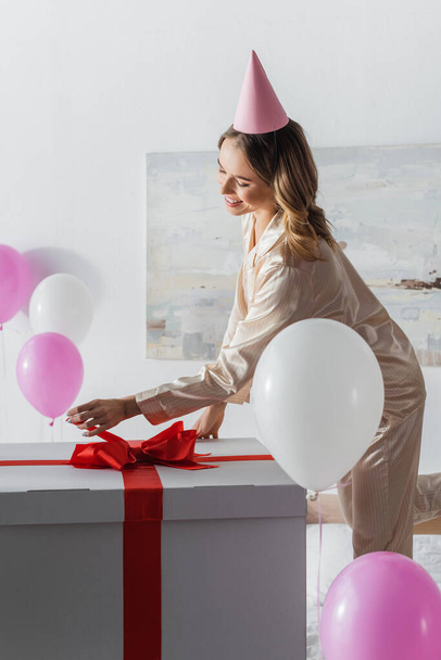 Smiling woman looking at big gift box near balloons in bedroom during birthday celebration  - Photo, Image