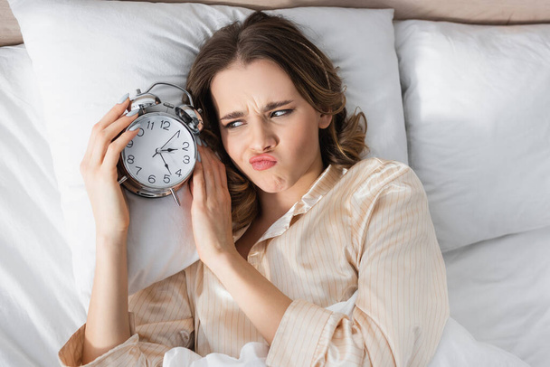 Top view of upset woman looking at alarm clock on bed  - Фото, изображение