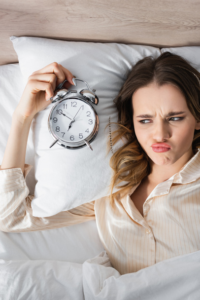 Top view of sad woman holding alarm clock on bed  - Foto, afbeelding
