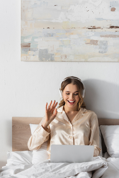 Mujer sonriente en auriculares con videollamada en el portátil en el dormitorio  - Foto, imagen