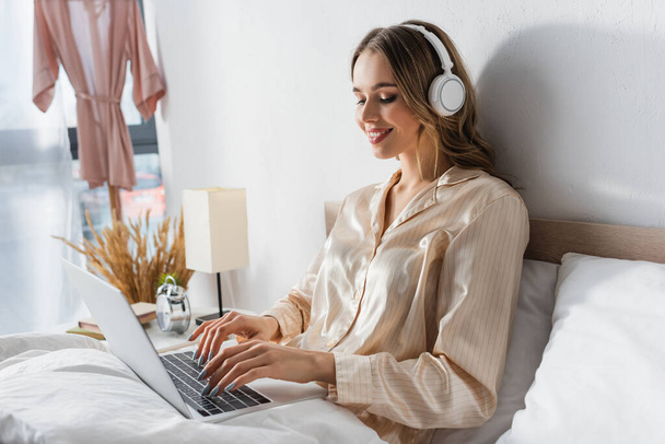 Smiling freelancer in headphones using laptop in bedroom  - Photo, Image