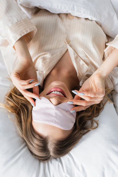 Вид сверху жизнерадостной женщины в маске для сна, лежащей на кровати  - Фото, изображение