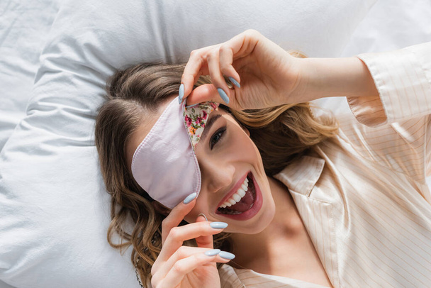 Top view of cheerful woman holding sleeping mask on bed  - Foto, immagini