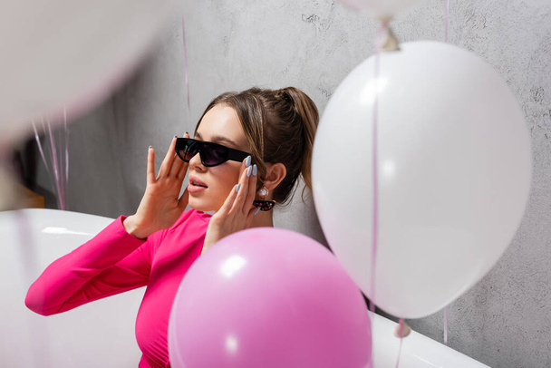 Young woman in sunglasses sitting in bathtub near blurred balloons on foreground  - Foto, imagen