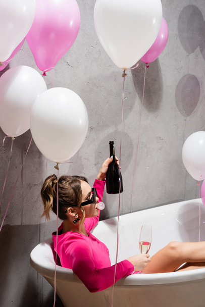 Woman in sunglasses blowing bubblegum and holding champagne in bathtub near balloons  - Φωτογραφία, εικόνα