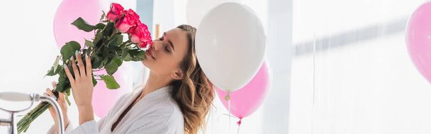 Woman in bathrobe smelling bouquet near balloons in bathroom, banner  - 写真・画像