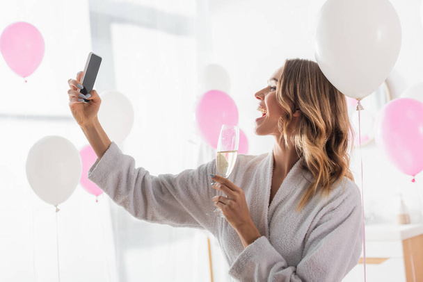 Side näkymä iloinen nainen ottaa selfie älypuhelimeen ja tilalla samppanjaa lähellä ilmapalloja  - Valokuva, kuva