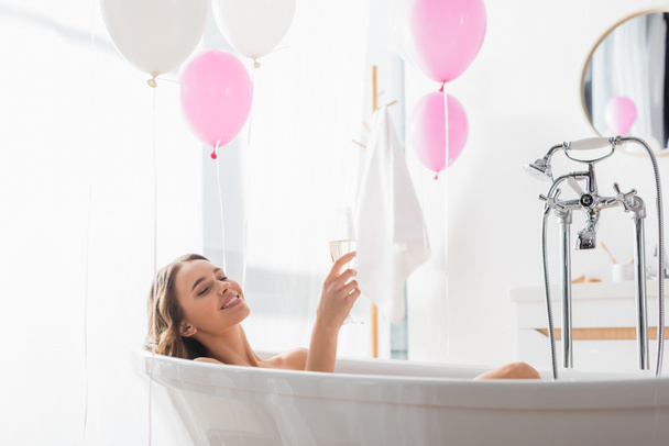 Glimlachende vrouw met champagne baden in de buurt ballonnen in de badkamer  - Foto, afbeelding