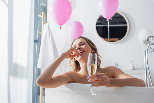 Bulanık şampanyalı olumlu bir kadın küvette balonların yanında oturuyor.  - Fotoğraf, Görsel
