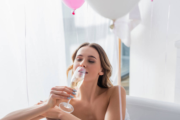 Woman drinking champagne while bathing near balloons at home  - Foto, Bild