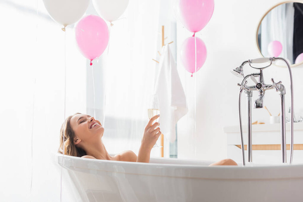 Happy woman holding glass of champagne near balloons in bathroom  - Фото, изображение
