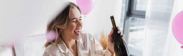 Cheerful woman holding bottle and glass of champagne near balloons in bedroom, banner  - Фото, изображение