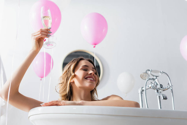 Vue en angle bas de la femme souriante avec verre de champagne assis dans la baignoire près des ballons festifs  - Photo, image