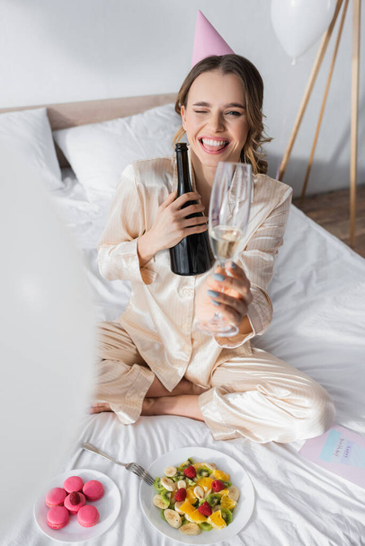 Woman in party cap holding champagne and winking at camera near fruit salad and macaroons on bed  - Foto, afbeelding