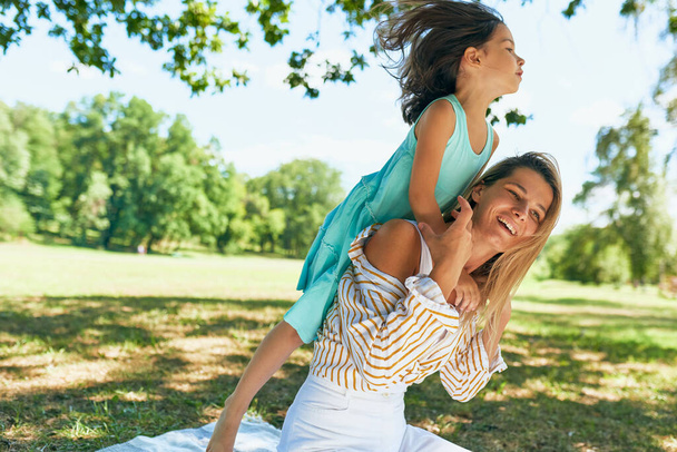 Горизонтальный образ счастливой любящей матери и ее милой дочери, играющей в парке во время пикника. Игривая красотка и маленькая девочка проводят время вместе в солнечный день. Мама и ребенок веселятся.. - Фото, изображение