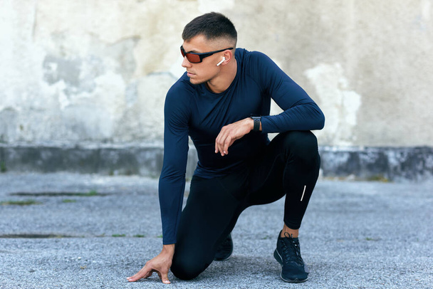 Horizontal image of a sporty male resting outdoors after the workout. Fitness man in blue sports clothing and eyewear exercising with earphones outside against the urban concrete wall. - Foto, Imagem